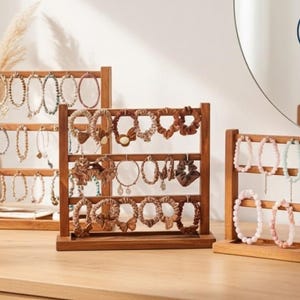 May include: Three wooden jewellery display stands showcasing bracelets and necklaces. The stands are made of light brown wood and have three horizontal bars for hanging jewellery. The jewellery includes beaded bracelets and necklaces in various colours.