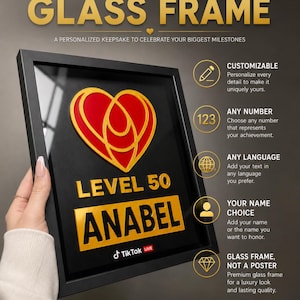 May include: A black framed glass art piece with gold text that reads "CUSTOM ACHIEVEMENT GLASS FRAME". The art features a red and gold heart design, the text "LEVEL 50 ANABEL", and the TikTok logo.