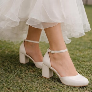 Puede incluir: Zapatos de tacón cuadrado color marfil con correas en el tobillo. Los zapatos tienen una punta redondeada y un pequeño cierre de hebilla. Los tacones miden aproximadamente 7.6 cm de alto. Los zapatos se usan sobre una superficie cubierta de hierba.