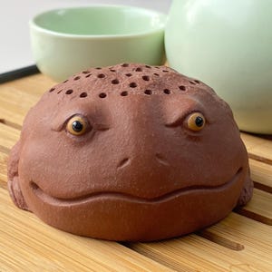 May include: A brown ceramic frog-shaped tea infuser with small holes on top, set on a bamboo tray. The infuser has yellow eyes and a smiling mouth. A light green tea cup and teapot are in the background.