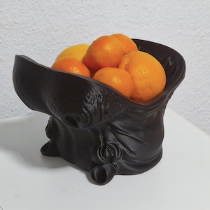 Könnte beinhalten: Eine schwarze, skulpturale Obstschale gefüllt mit Orangen und einer Zitrone. Die Schale hat ein einzigartiges, drapiertes Design, das einer Stofftasche ähnelt. Die Orangen sind leuchtend orange und kontrastieren mit der dunklen Schale. Die Schale steht auf einer weißen Oberfläche.