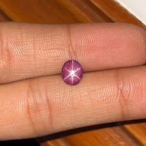 May include: A small, round, cabochon-cut star ruby gemstone with a deep magenta-pink hue. The stone displays a distinct, centered six-rayed star effect, known as asterism, which appears as a bright white light reflection across the polished surface.