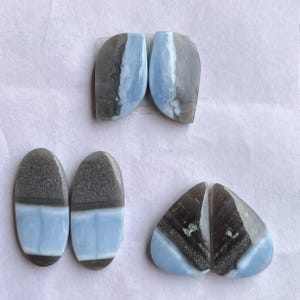 Puede incluir: Una colección de gemas pulidas en varias formas. Las piedras presentan una combinación de colores gris, blanco y azul claro. Las formas incluyen óvalos, lágrimas y formas irregulares, mostrando patrones naturales.