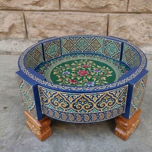 May include: A round, hand-painted wooden tray with a blue exterior and intricate floral and paisley designs. The interior features a green background with a floral pattern. The tray is supported by four small, wooden legs.