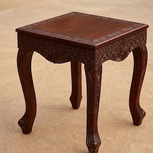 Pode incluir: Uma mesa lateral quadrada marrom escuro com entalhes ornamentados. A mesa tem um design floral detalhado ao redor das bordas do tampo e ao longo das pernas curvas. A mesa tem aproximadamente 51 cm de altura.
