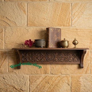 May include: A dark brown wooden shelf with carved floral patterns. It displays a book, small decorative containers, and a flower against a textured stone wall. The text "Wood wala shop" is visible.