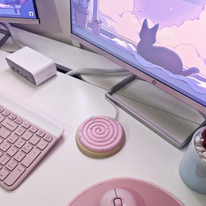 Peut inclure: Tapis de souris rose et beige en forme de biscuit avec un motif en spirale et des vermicelles. Un clavier, une souris et un adaptateur secteur blanc sont également sur le bureau blanc. Un écran d'ordinateur affiche une silhouette de chat.