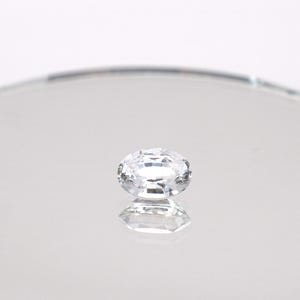 May include: A clear, faceted gemstone with a rectangular shape, set on a reflective, circular surface. The gemstone is positioned in the centre, reflecting its form. The background is a soft, white gradient.