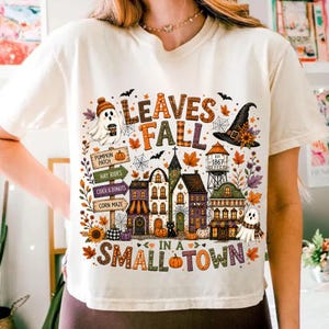 May include: A cream-colored t-shirt featuring a colorful autumn-themed graphic with the text "LEAVES FALL IN A SMALL TOWN". The design includes a ghost, a witch hat, a water tower, houses, pumpkins, fall foliage, and signs for a pumpkin patch.