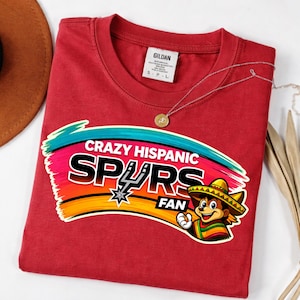 Op de afbeelding: Een rood t-shirt met de tekst "Crazy Hispanic Spurs Fan" in een kleurrijk ontwerp. De afbeelding bevat een cartoonfiguur met een sombrero en het Spurs-logo. Een gouden ketting is over het shirt gedrapeerd.