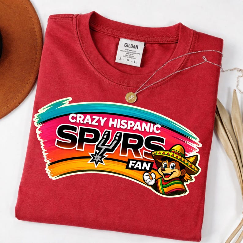 Op de afbeelding: Een rood t-shirt met de tekst "Crazy Hispanic Spurs Fan" in een kleurrijk ontwerp. De afbeelding bevat een cartoonfiguur met een sombrero en het Spurs-logo. Een gouden ketting is over het shirt gedrapeerd.