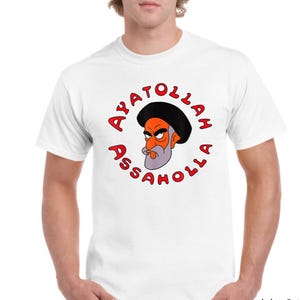 Puede incluir: Camiseta blanca con una ilustración de dibujos animados de un hombre con turbante negro, cara naranja y barba gris. El texto rojo "AYATOLLAH ASSHOLLA" está impreso en círculo alrededor de la imagen.