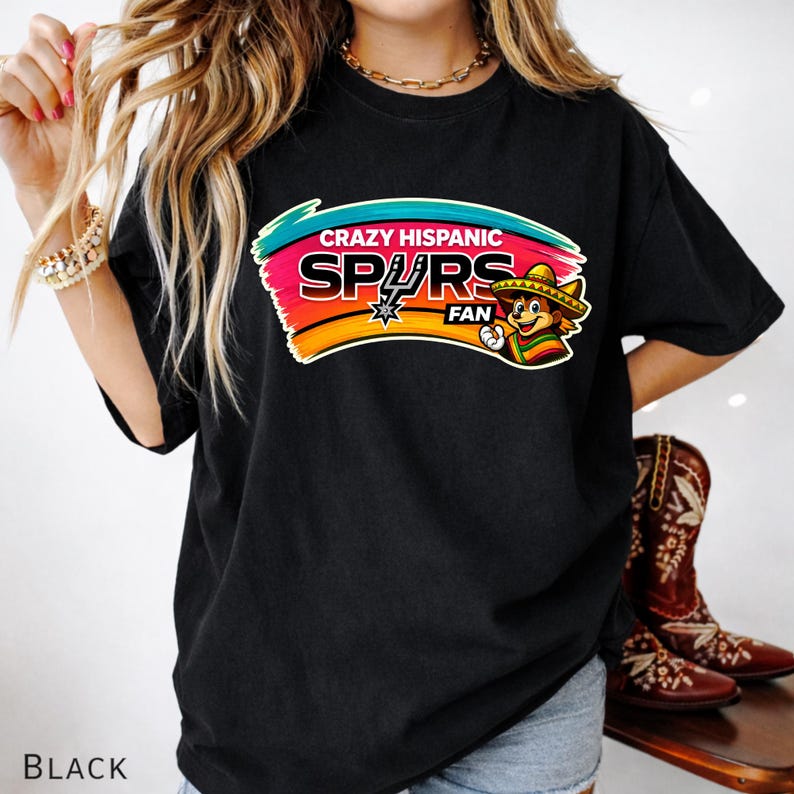Op de afbeelding: Zwart t-shirt met een kleurrijke afbeelding met de tekst "Crazy Hispanic Spurs Fan". De afbeelding toont het Spurs-logo, een stripfiguur met een sombrero en een levendig kleurenschema van teal, roze, oranje en geel.