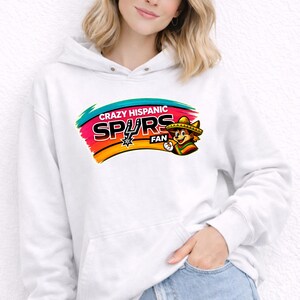Op de afbeelding: Witte hoodie met een kleurrijke afbeelding met de tekst "Crazy Hispanic Spurs Fan". De afbeelding toont een cartoonfiguur met een sombrero en een teamlogo. De hoodie heeft een voorzak en een capuchon met trekkoord.