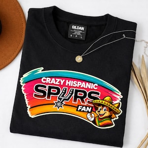 Op de afbeelding: Zwart sweatshirt met de tekst "CRAZY HISPANIC SPURS FAN" in een kleurrijk ontwerp. De afbeelding bevat een cartoonfiguur met een sombrero en het Spurs-logo. Een gouden ketting is ook zichtbaar.