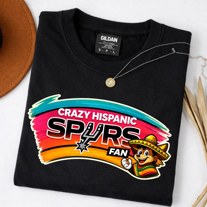 Op de afbeelding: Zwart sweatshirt met de tekst "CRAZY HISPANIC SPURS FAN" in een kleurrijk ontwerp. De afbeelding bevat een cartoonfiguur met een sombrero en het Spurs-logo. Een gouden ketting is ook zichtbaar.