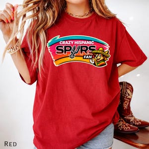 Op de afbeelding: Rood t-shirt met een kleurrijke afbeelding met de tekst "Crazy Hispanic Spurs Fan". De afbeelding toont een cartoonfiguur met een sombrero. Het woord "RED" staat linksonder op het shirt.