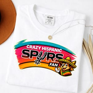 Op de afbeelding: Wit T-shirt met een kleurrijke afbeelding met de tekst "Crazy Hispanic Spurs Fan". Het ontwerp bevat het Spurs-logo en een cartoonfiguur met een sombrero en een serape. Het shirt heeft een ronde hals en korte mouwen.