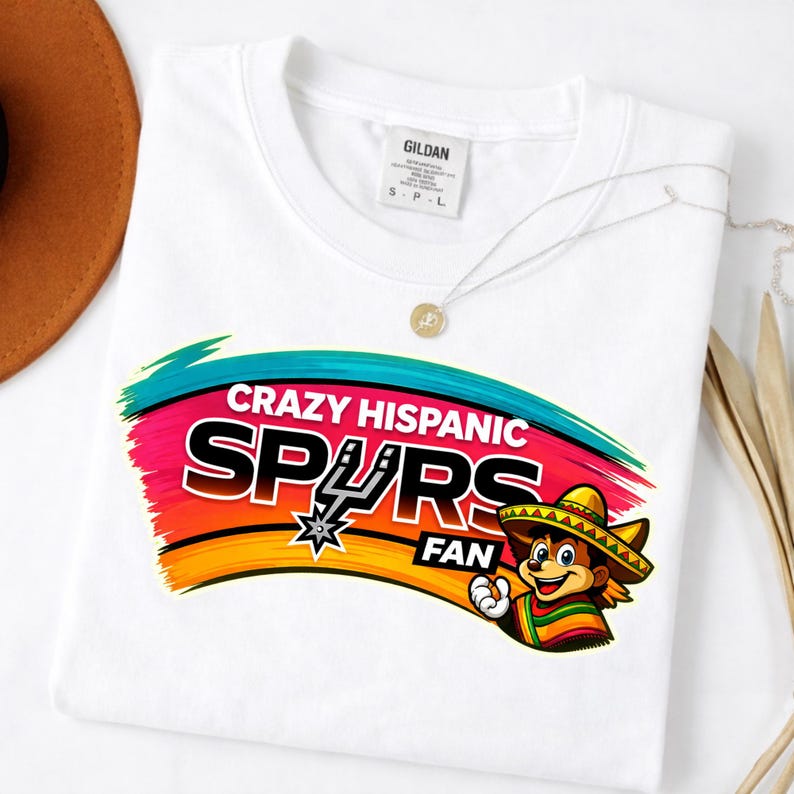 Op de afbeelding: Wit T-shirt met een kleurrijke afbeelding met de tekst "Crazy Hispanic Spurs Fan". Het ontwerp bevat het Spurs-logo en een cartoonfiguur met een sombrero en een serape. Het shirt heeft een ronde hals en korte mouwen.