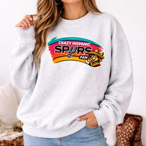 Op de afbeelding: Een lichtgrijs sweatshirt met de tekst "CRAZY HISPANIC SPURS FAN" in een kleurrijk ontwerp. Het ontwerp bevat een cartoonfiguur met een sombrero en een Spurs-logo.
