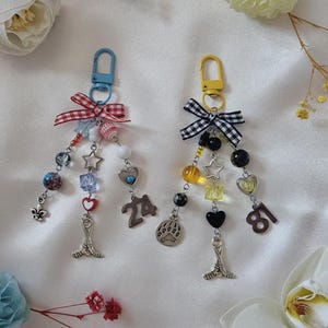 Peut inclure: Deux porte-clés décoratifs avec des fermoirs mousquetons en métal, des nœuds en vichy et des perles assorties. Le porte-clés gauche a un fermoir bleu, un nœud vichy rouge et le chiffre 24. Le droit a un fermoir jaune, un nœud vichy noir et le chiffre 81.