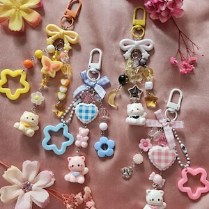 Op de afbeelding: Vier decoratieve sleutelhangers met kattenfiguurtjes, strikjes en diverse bedels. De sleutelhangers hebben gele, blauwe en roze kleurenschema's met ster-, hart- en bloemvormen, bevestigd aan metalen karabijnsluitingen en lintjes.