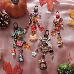 May include: Four decorative keychains featuring autumn-themed charms including a raccoon, a fox face, a highland cow, and a squirrel. Each keychain includes a fabric bow, colorful beads, and miniature charms like mushrooms, hearts, and leaves.