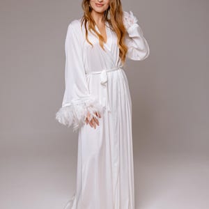 May include: A long, white satin robe with a wrap-around design and a matching belt. The sleeves feature feather trim at the cuffs. The robe is floor-length and has a flowing silhouette.
