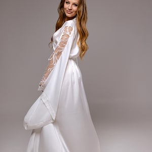May include: A floor-length white satin robe featuring long, wide sleeves with intricate white lace panels along the upper arms. The garment includes a matching fabric tie belt at the waist and a smooth, flowing silhouette that reaches the floor.