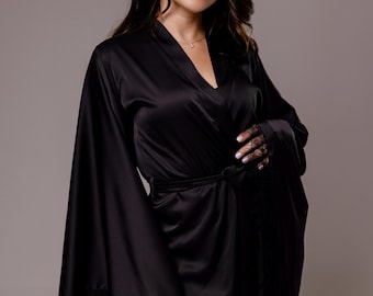 May include: A black satin robe featuring wide, flowing sleeves and delicate black lace trim along the sleeve edges and hem. The garment includes a matching fabric tie belt at the waist and a V-neckline, crafted from a smooth, reflective material.