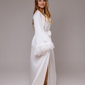 Może przedstawiać: Biały satynowy szlafrok z długimi rękawami i pasującym paskiem. Rękawy są obszyte białymi piórami. Szlafrok jest otwarty z przodu, odsłaniając złote sandały. Modelka się uśmiecha.