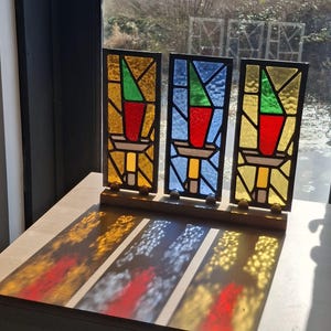May include: Three stained glass panels in a row, each with a different colored background: yellow, blue, and yellow. Each panel features a red vase-like shape with a green triangle on top. The panels are set on a wooden base.