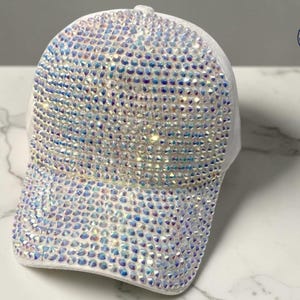 May include: White baseball cap with iridescent rhinestones covering the front and peak. The cap has a curved peak and a rounded crown. The rhinestones reflect a rainbow of colours. A logo is visible on the side.