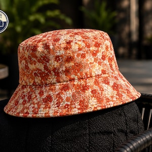 May include: A floral bucket hat in shades of orange, red, and cream. The hat features a repeating pattern of small flowers. The brim is slightly angled, and the hat is sitting on a black surface. The logo "Stylish Caps" is in the upper left corner.