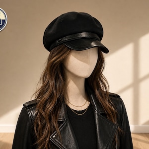 May include: A black newsboy cap with a leather visor is displayed on a mannequin. The cap has a rounded crown and a structured brim. The mannequin is wearing a black leather jacket and a gold necklace. The logo "Stylish Caps" is in the upper left corner.
