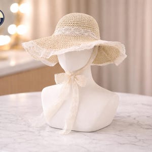 May include: A straw sun hat with a wide brim and a delicate lace trim. The hat is displayed on a white mannequin head, with a matching ribbon tied in a bow under the chin. The hat is a light beige color.