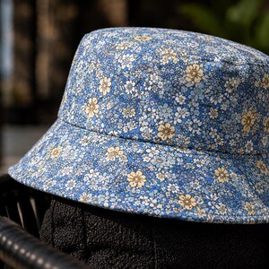 May include: A blue bucket hat with a floral pattern. The hat features a light blue background with small white and yellow flowers. The brim is slightly angled, and the hat is made of a soft fabric. This hat is a stylish accessory.