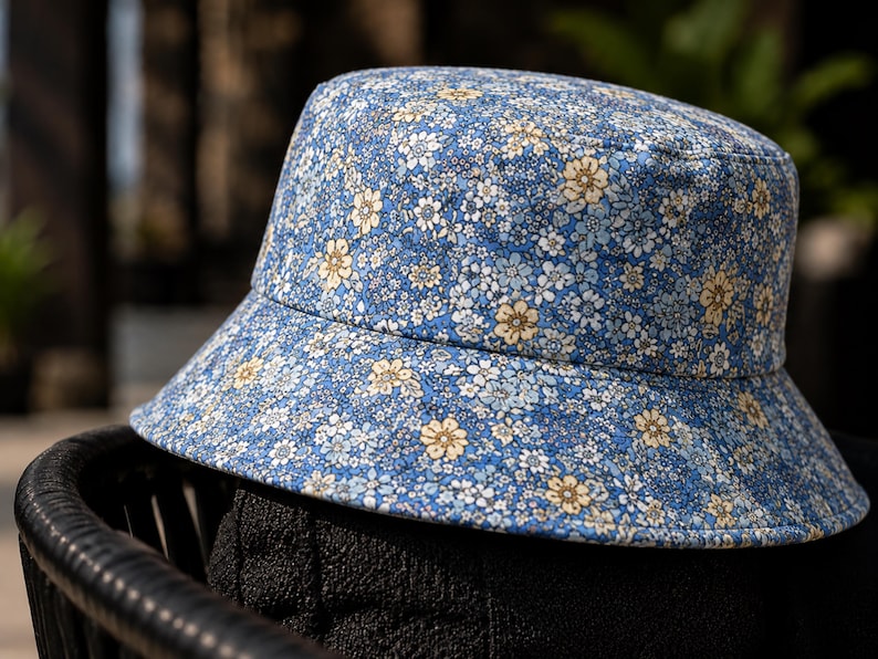 May include: A blue bucket hat with a floral pattern. The hat features a light blue background with small white and yellow flowers. The brim is slightly angled, and the hat is made of a soft fabric. This hat is a stylish accessory.