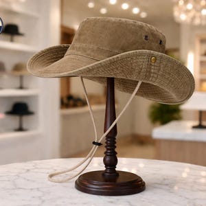 Op de afbeelding: Een bruinbeige safarihoed met een brede rand en kinband wordt getoond op een donkerhouten hoedenstandaard. De hoed heeft een licht verweerde uitstraling en is gemaakt van een textuurstof. De hoedenstandaard staat op een marmeren tafel.