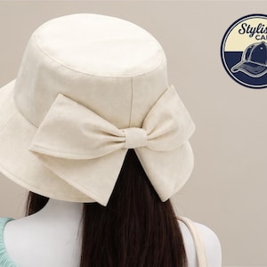 May include: A cream-coloured bucket hat with a large bow on the back. The hat has a wide brim and a rounded crown. The image also includes a logo that says "Stylish Caps" with a drawing of a baseball cap.