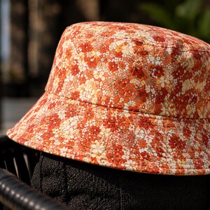 May include: A floral bucket hat in shades of orange, red, and white. The hat features a wide brim and is covered in a repeating pattern of small flowers. The hat is sitting on a black surface.