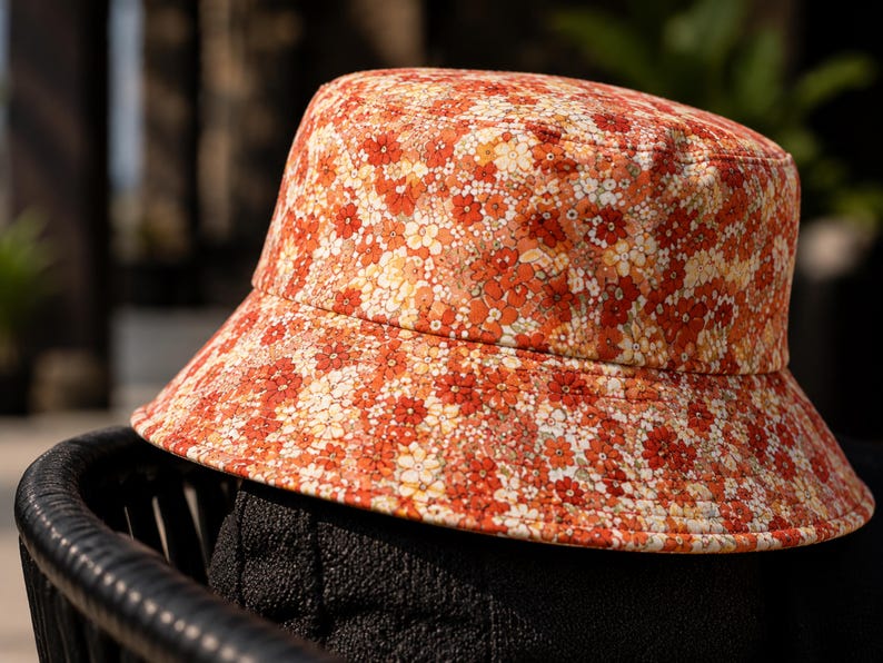 May include: A floral bucket hat in shades of orange, red, and white. The hat features a wide brim and is covered in a repeating pattern of small flowers. The hat is sitting on a black surface.