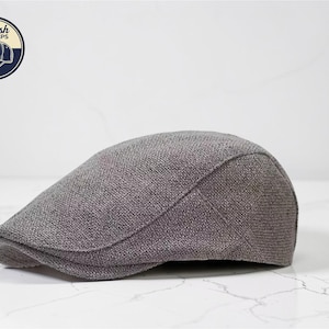 May include: A gray, textured flat cap, also known as a newsboy cap, sits on a white marble surface. The cap has a rounded shape and a small, stiff brim. A logo in a circle with the words "Stylish Caps" and a cap illustration is in the upper left corner.