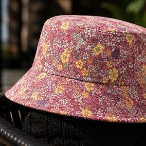 May include: A pink bucket hat with a floral pattern featuring yellow, white, and purple flowers. The hat is made of fabric and has a wide brim. The hat is sitting on a black surface.