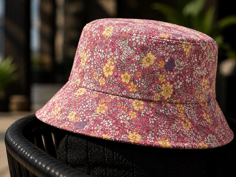 May include: A pink bucket hat with a floral pattern featuring yellow, white, and purple flowers. The hat is made of fabric and has a wide brim. The hat is sitting on a black surface.