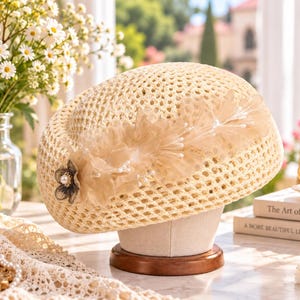 Puede incluir: Una boina de ganchillo beige con un patrón de tejido abierto, adornada con un acento decorativo lateral que consiste en pétalos de tela en capas, hojas de malla transparente y una pequeña flor central con una perla artificial.