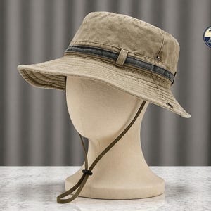 Puede incluir: Un sombrero de pescador de color caqui con una banda azul oscuro y una correa de barbilla ajustable. El sombrero se exhibe en una cabeza de maniquí. El sombrero tiene un ala ancha y está hecho de una tela texturizada. El sombrero tiene un logotipo "Stylish Caps".