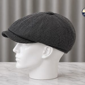 Könnte beinhalten: Dunkelgraue Newsboy-Cap mit Fischgrätenmuster und kurzem Schirm. Die Kappe wird auf einem weißen Schaufensterkopf präsentiert. Sie hat einen klassischen, Vintage-Stil. Das Logo "Stylish Caps" ist oben rechts sichtbar.