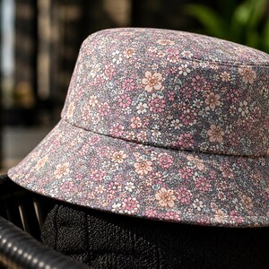 May include: A floral print bucket hat with a gray background and pink and white flowers. The hat is sitting on a black chair. The hat has a wide brim and a rounded crown.