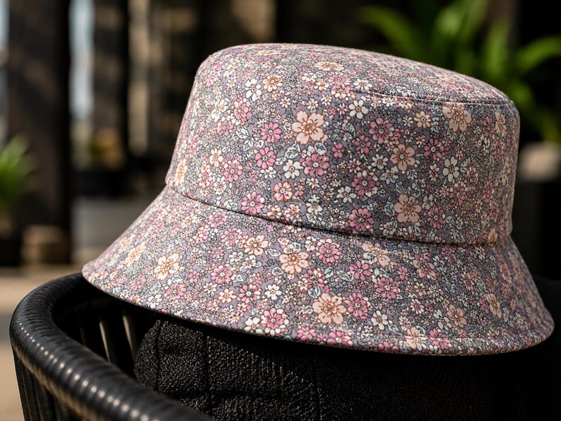 May include: A floral print bucket hat with a gray background and pink and white flowers. The hat is sitting on a black chair. The hat has a wide brim and a rounded crown.